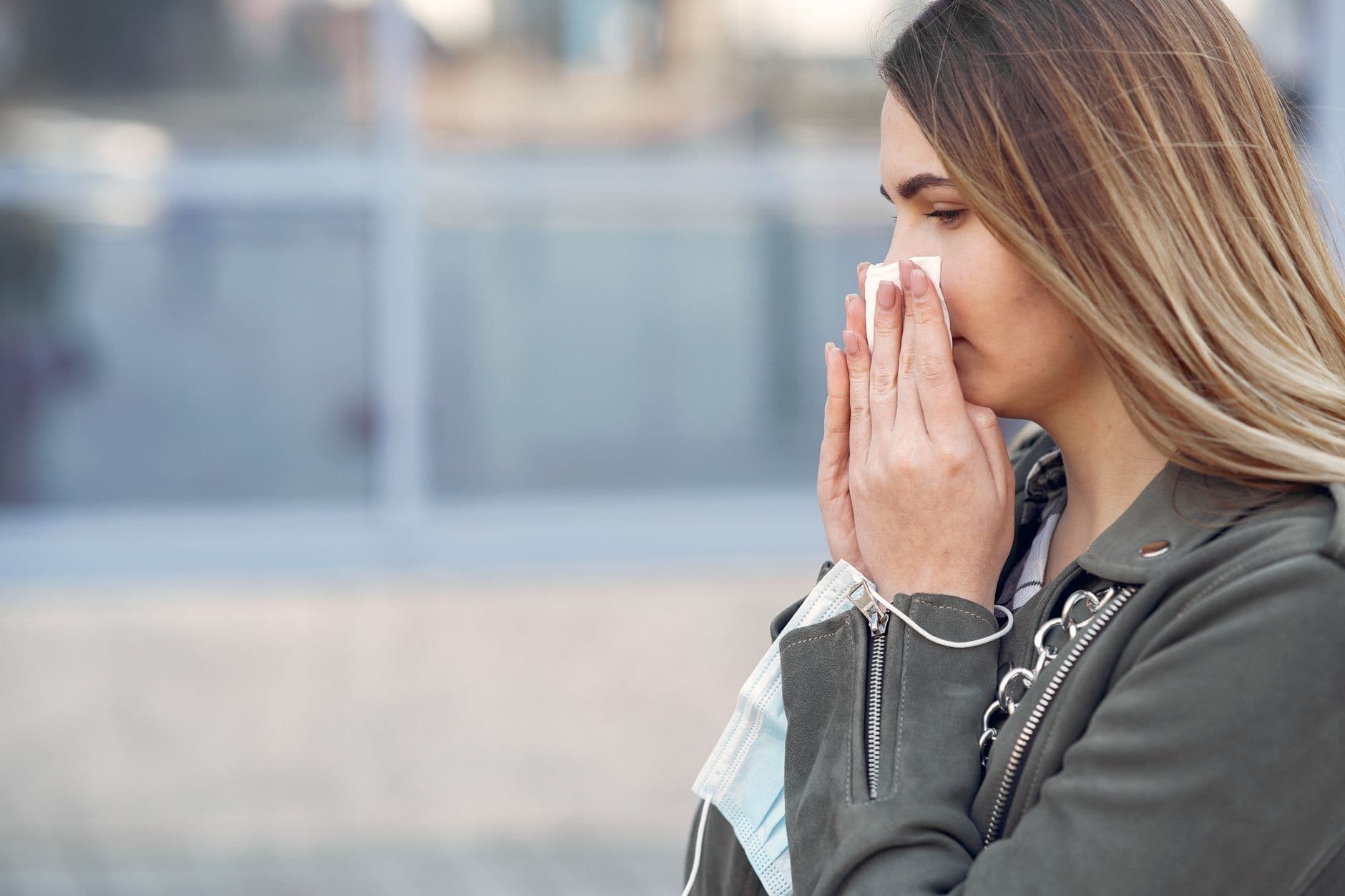 A woman sneezes outdoors while holding a tissue and a mask, reflecting health concerns.