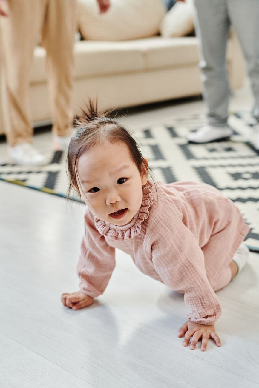 A cute East Asian baby girl crawling on a living room floor with family members in the background.