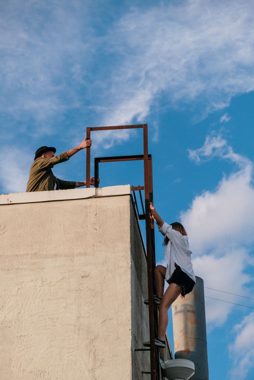 Two people climbing a rooftop ladder under a clear blue sky, embodying urban exploration and adventure.