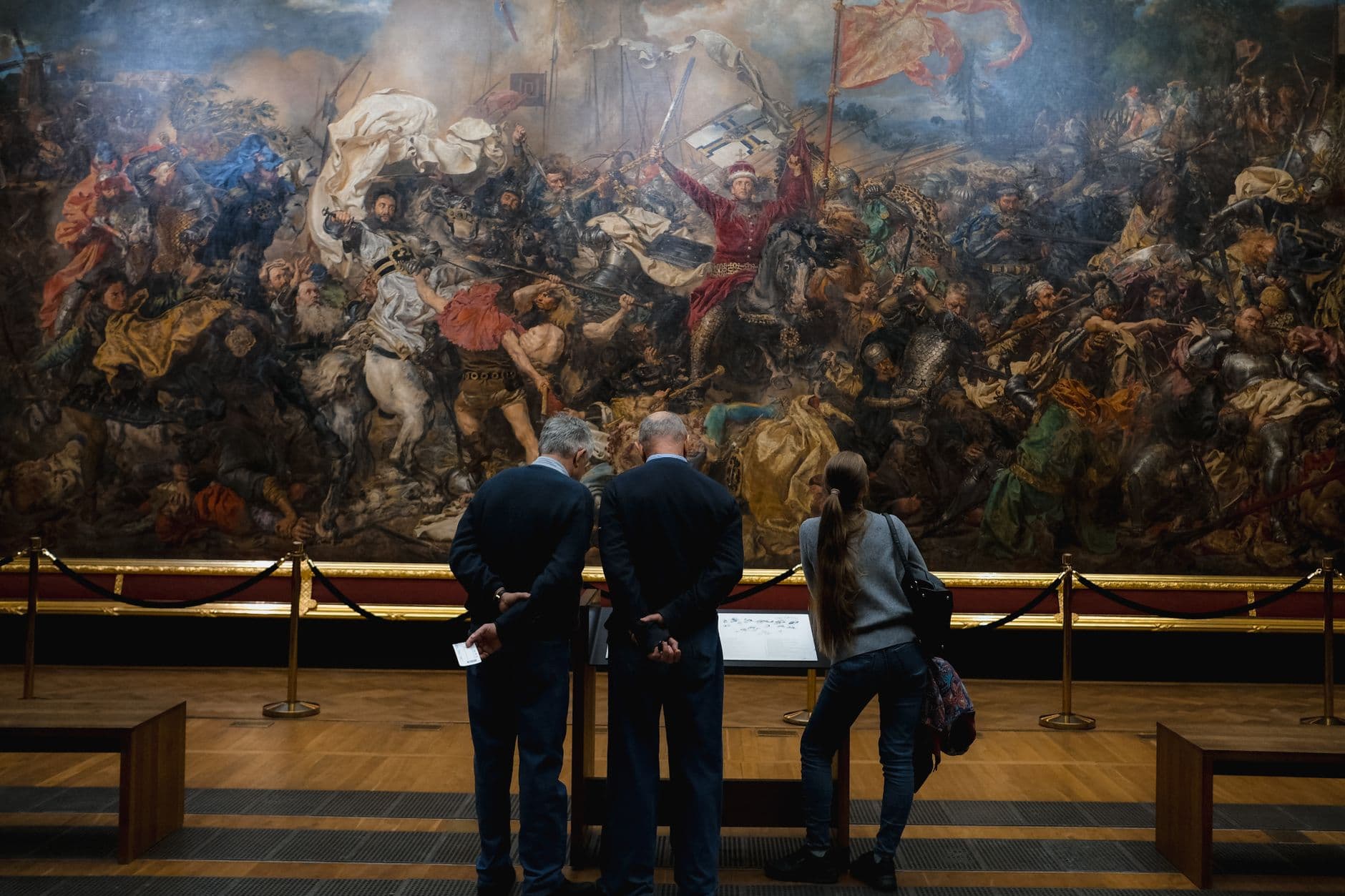 People viewing a large, detailed historical battle painting in a museum setting.
