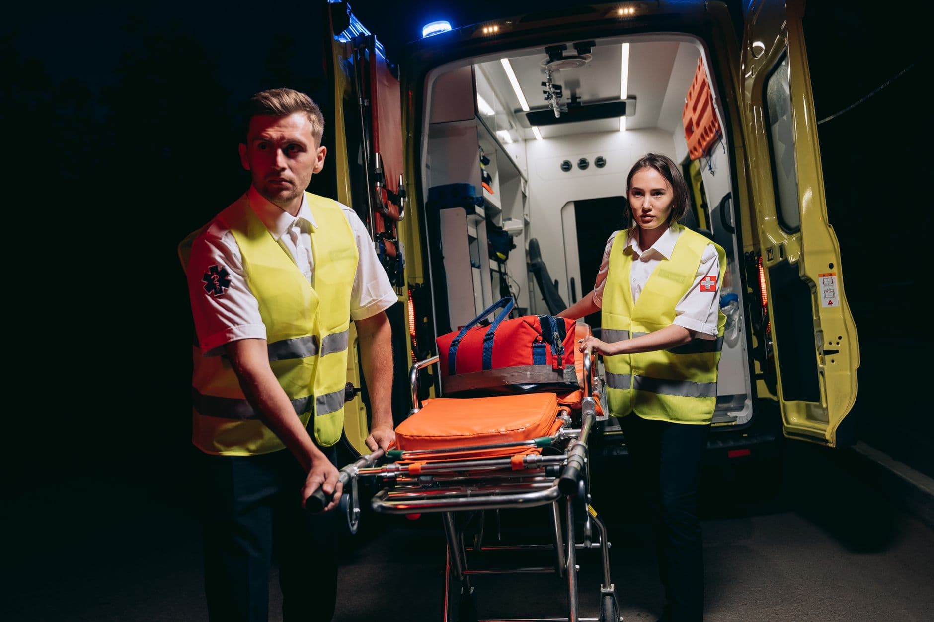 Emergency responders in high-visibility vests prepare a stretcher by an ambulance at night.
