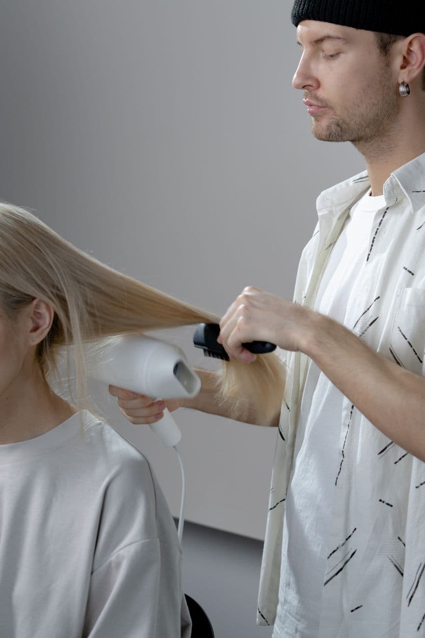 Hairstylist uses blow dryer and brush to style client's long blonde hair in salon.