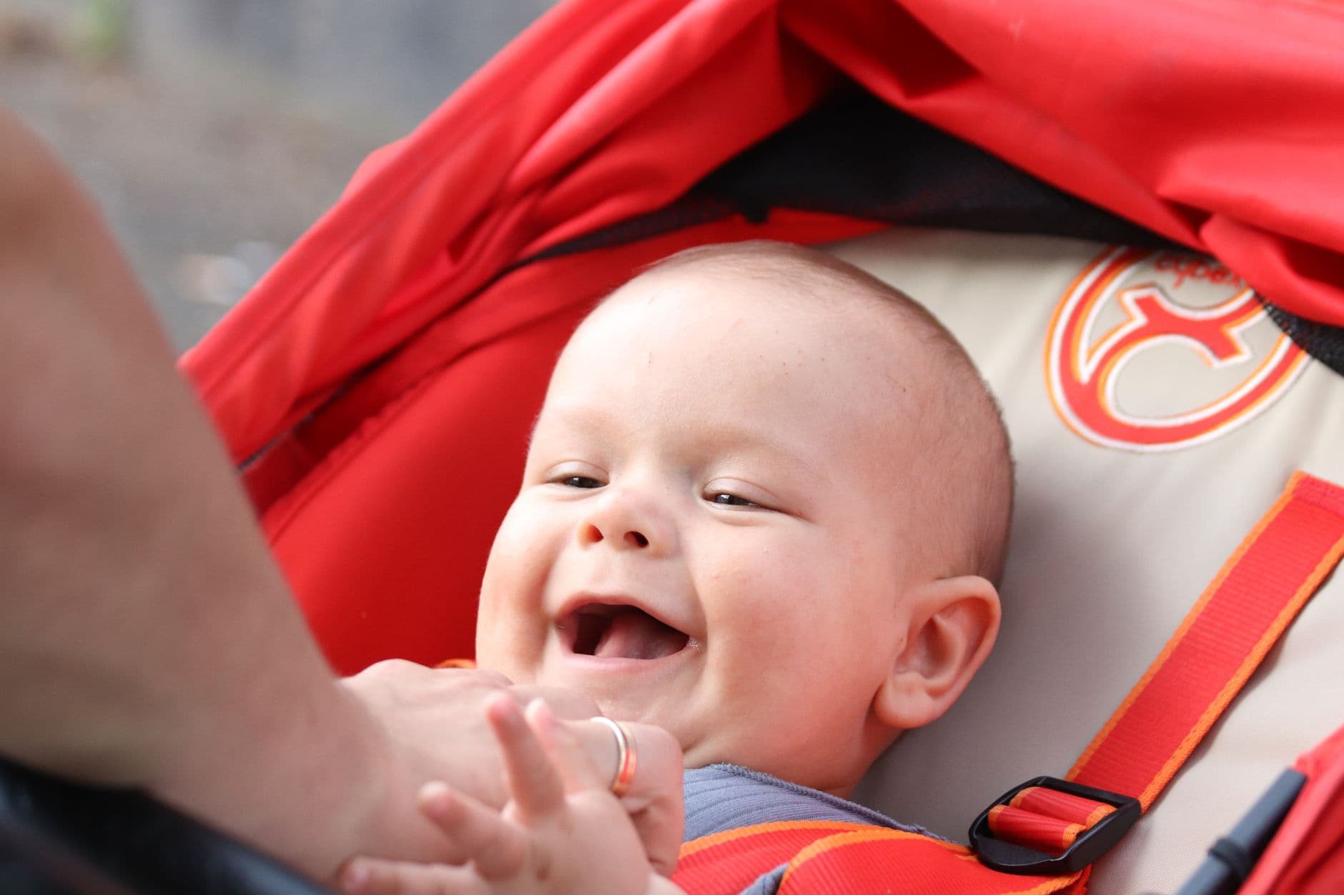 Adorable baby laughing joyfully while lying in a red stroller outside.
