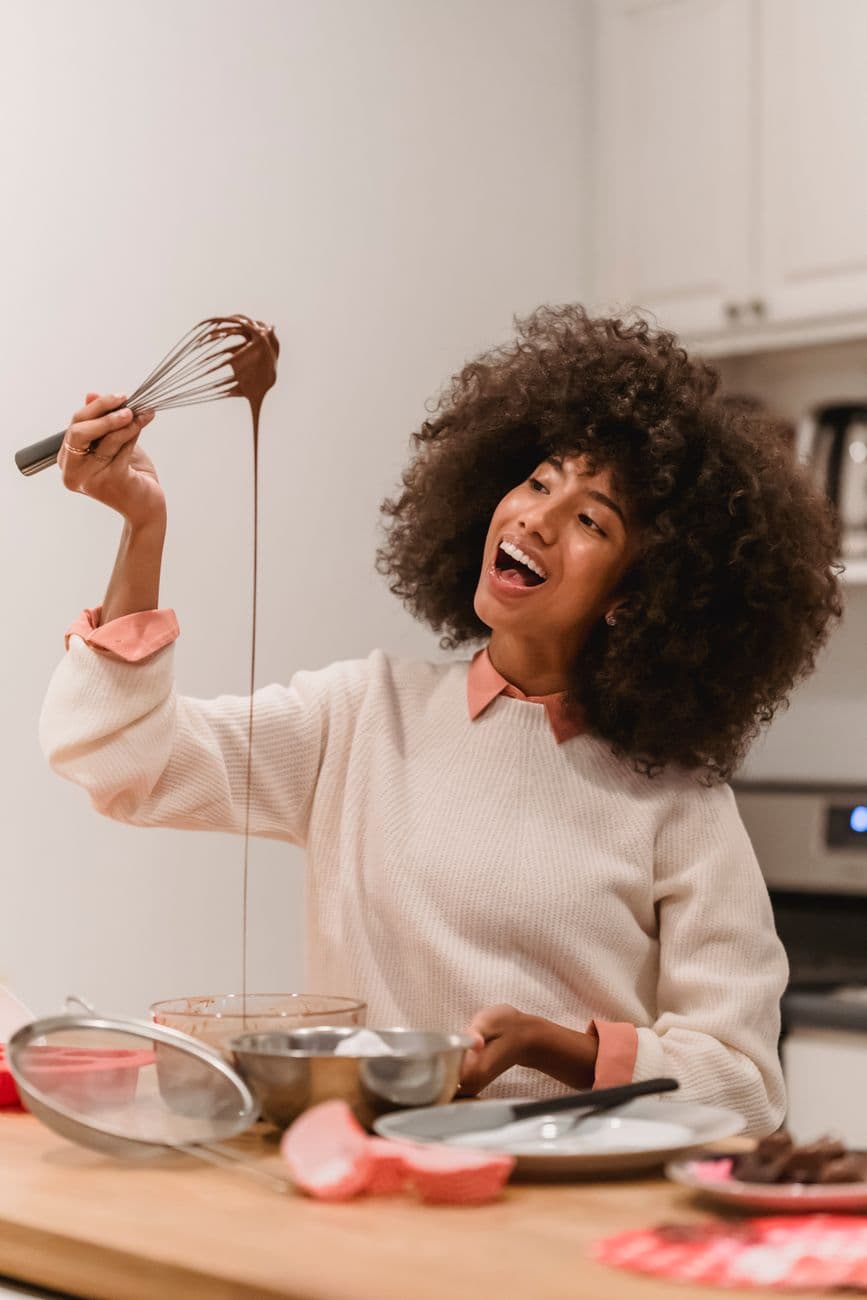 Joyful African American female cook looking at whisk with flowing batter while standing at table with various kitchenware at home