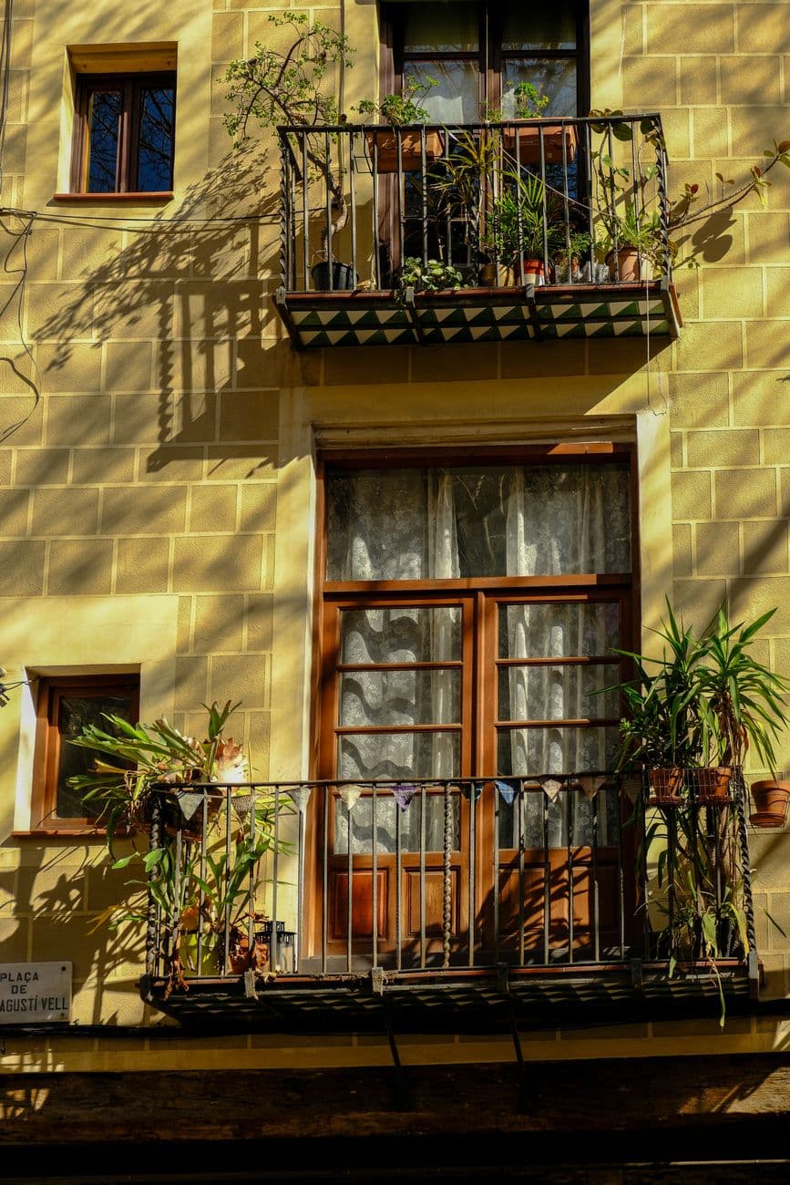 Sunny facade of a Barcelona townhouse featuring lush balcony plants and wooden windows.