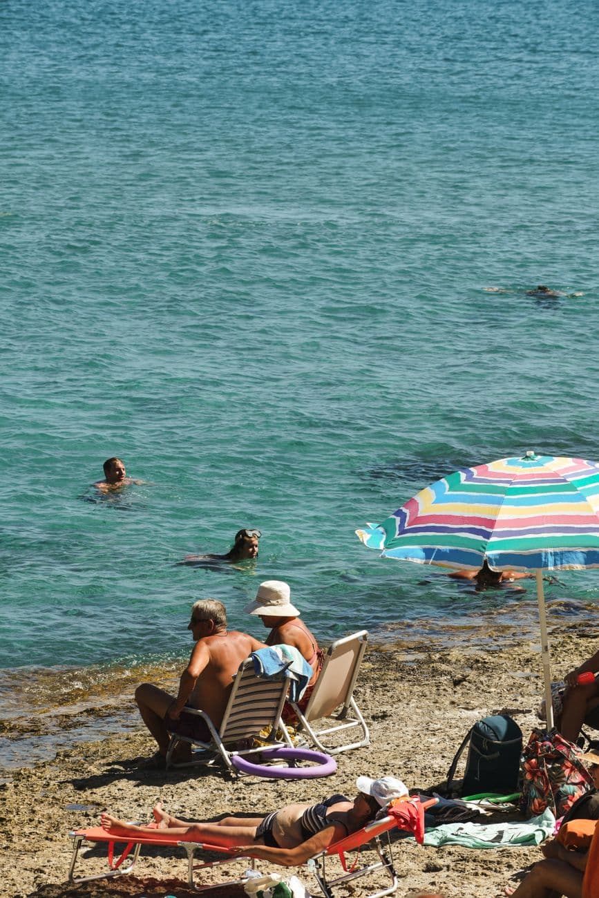People enjoying a sunny day at a Greek beach with calm waters and a vibrant umbrella.