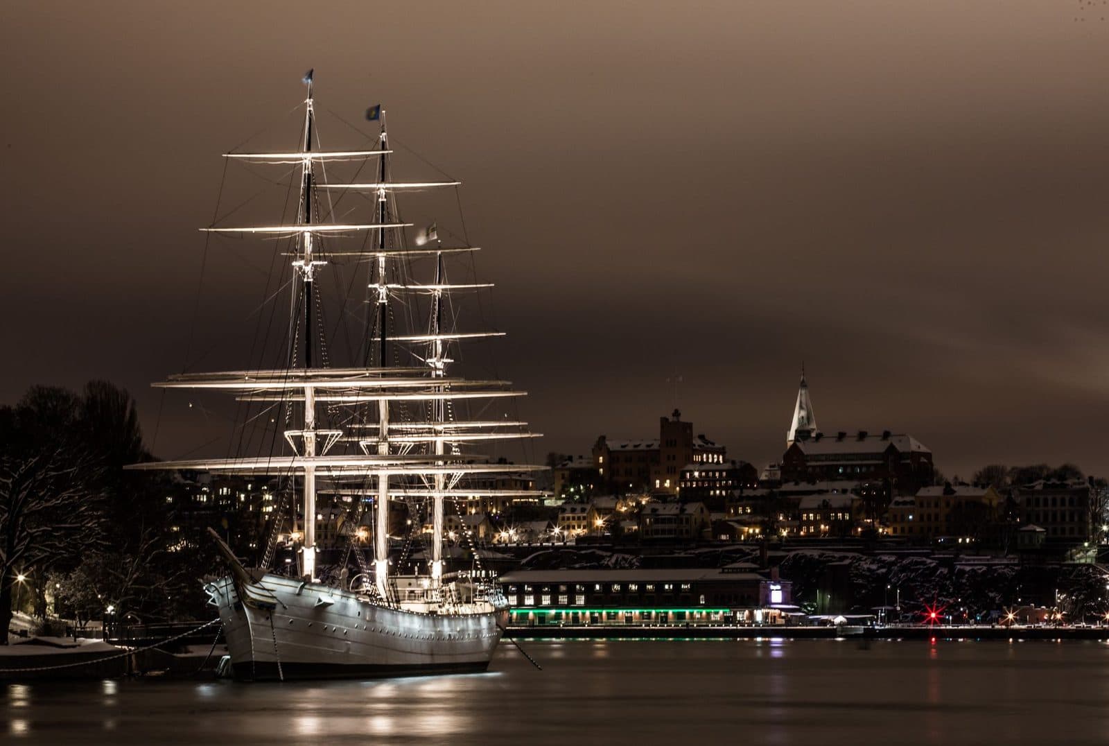 A majestic illuminated sailboat moored in Stockholm's harbor at night, showcasing beautiful city lights.