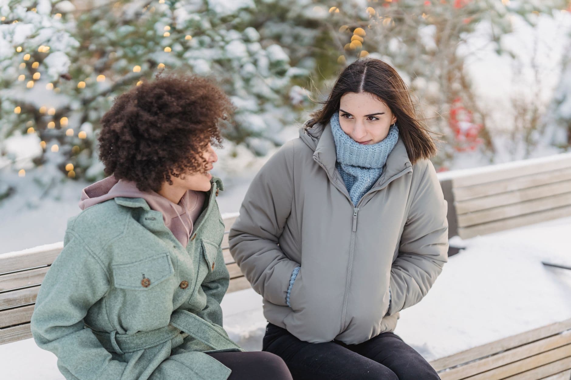 High angle of multiethnic girlfriends sitting with hands in pockets on bench and having conversation in cold winter day