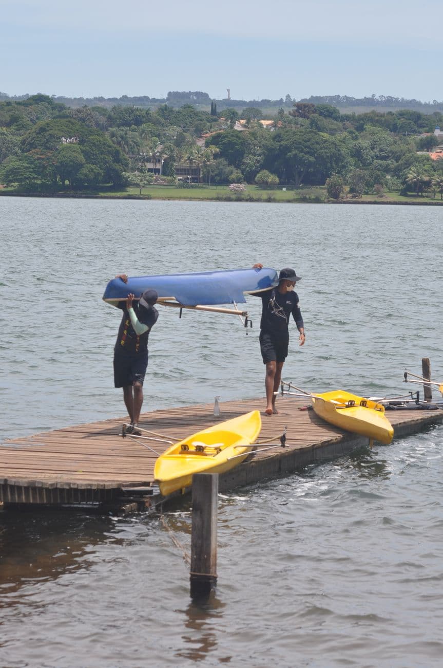 Two men carrying a blue canoe onto a dock with kayaks in Brasília, Brazil.
