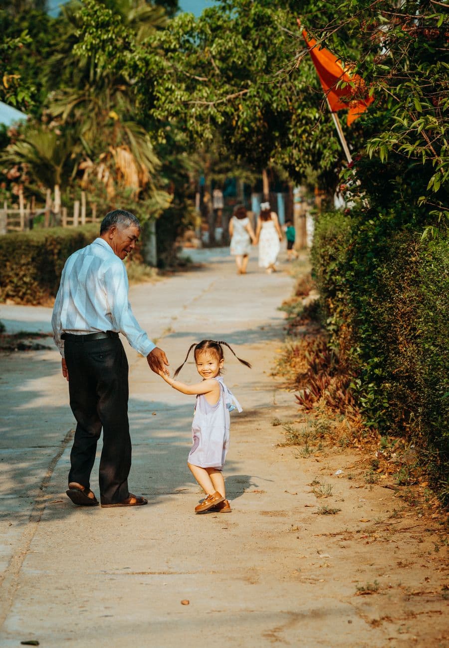 Elderly man and child walk hand-in-hand on a sunny street in Da Nang, Vietnam, surrounded by greenery.