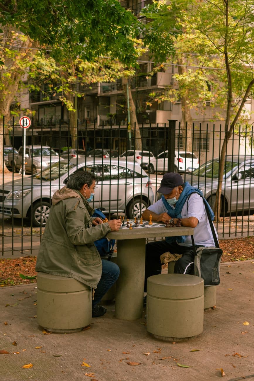 Two men in masks playing chess at a park table surrounded by parked cars and trees.