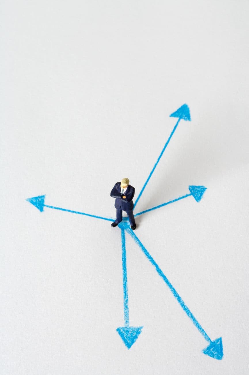 Miniature businessman stands at crossroads of blue arrows on white background, symbolizing choices.