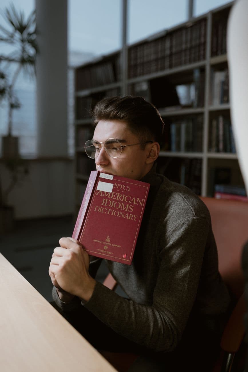Young man wearing glasses, holding an American idioms dictionary in a peaceful library setting.