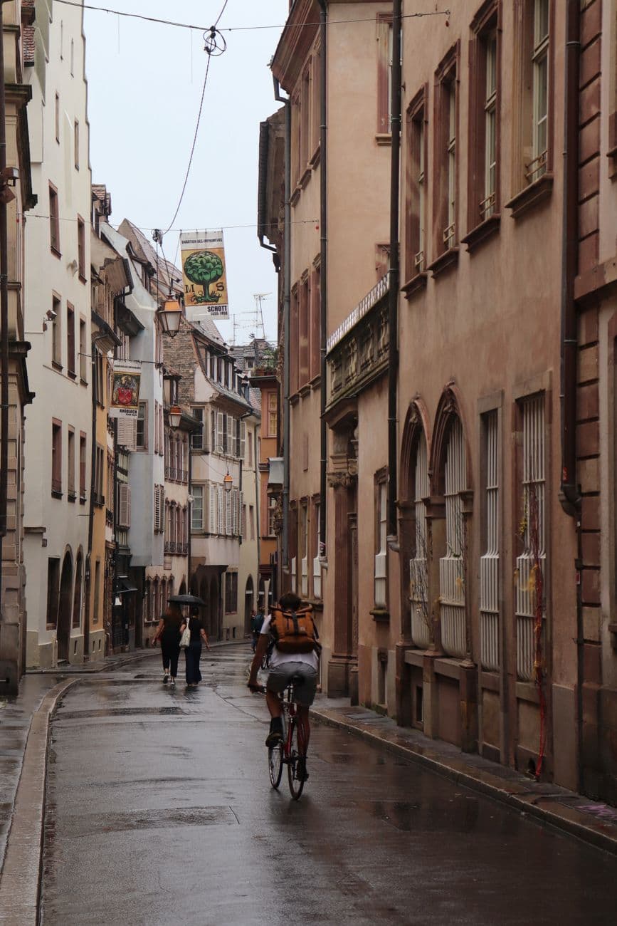 Explore a quaint street in Strasbourg, Grand Est, featuring historic architecture and a cyclist on a rainy day.