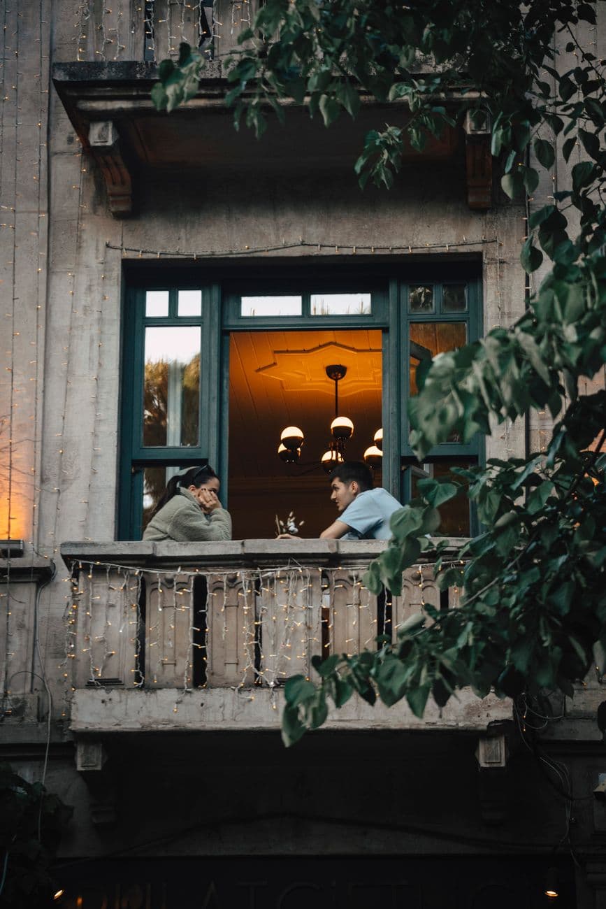 Two people enjoy a candid conversation on a balcony adorned with string lights, offering a warm, inviting ambiance.