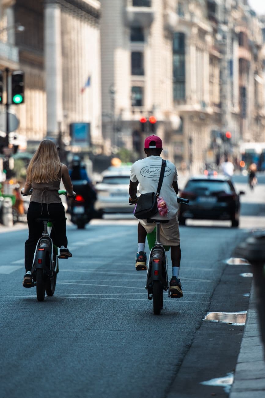 Two adults riding bicycles down a bustling Paris street on a sunny day.