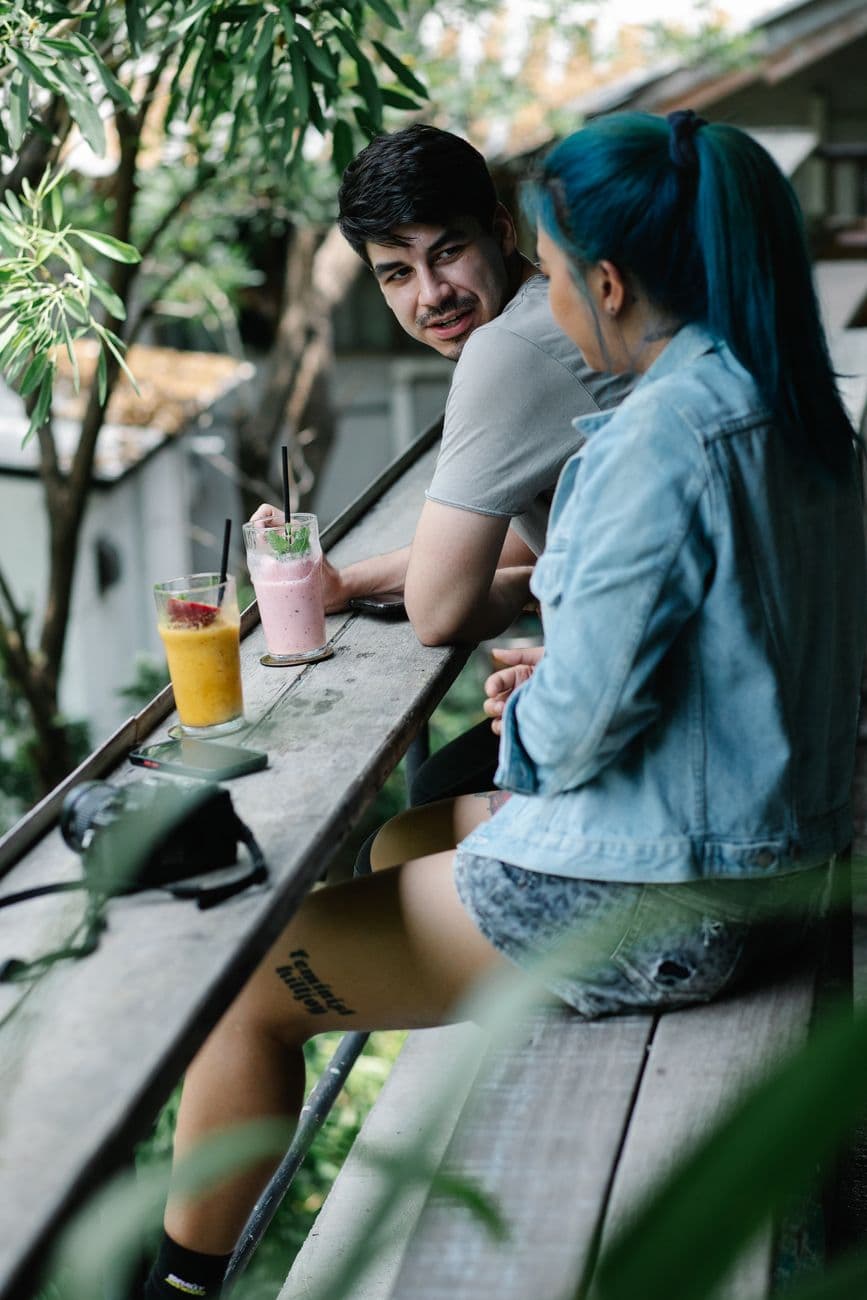 Couple of tourists sitting at wooden table with refreshing drinks on veranda of cafeteria near tall green trees on summer day
