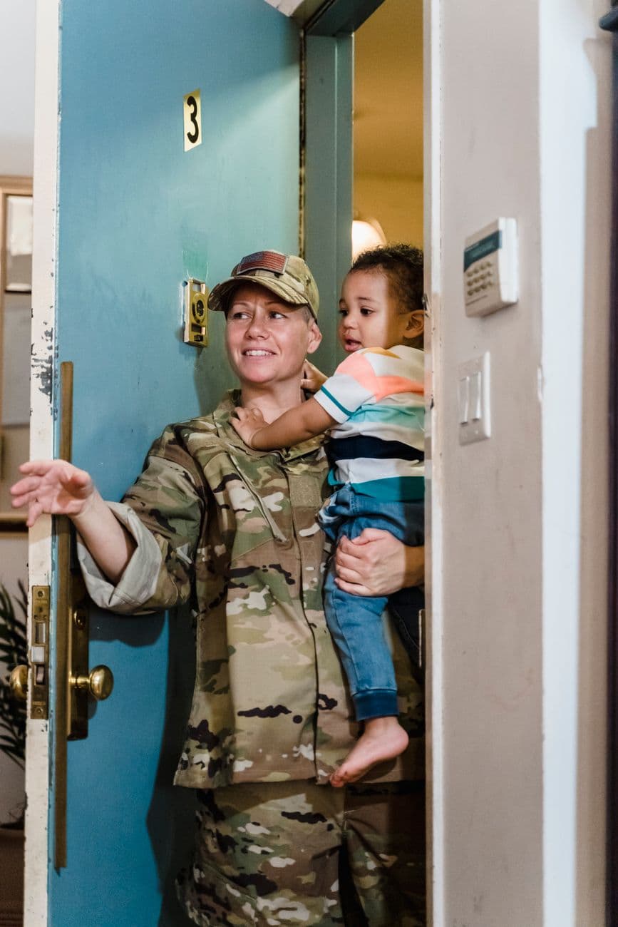 Smiling military woman in uniform welcomes home toddler child, standing in doorway.