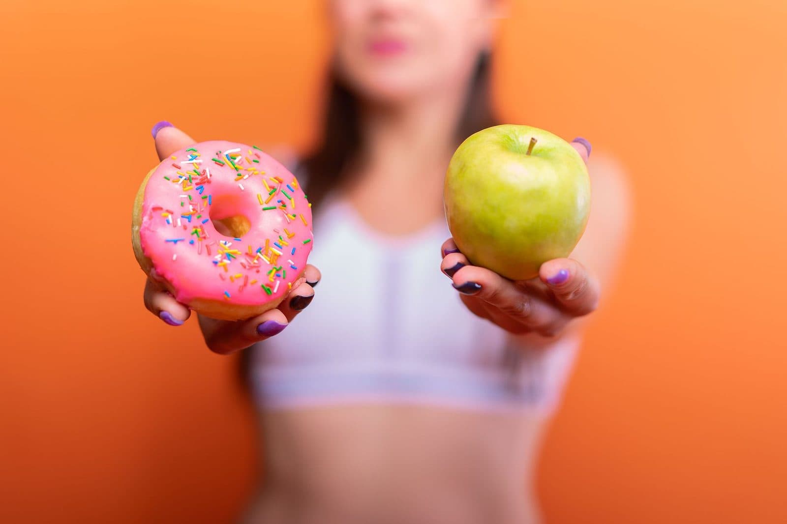 A woman holds a green apple and a pink donut with sprinkles against an orange background, emphasizing choice.