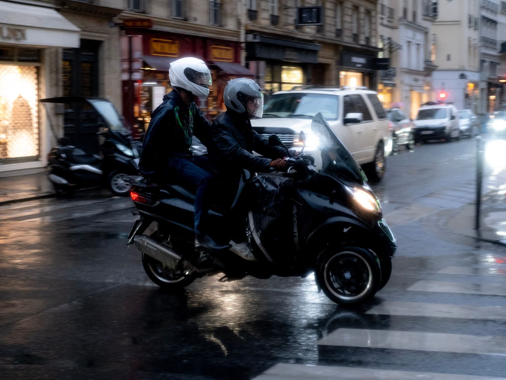 Two motorcyclists traverse a rainy city street at night, highlighting urban travel.