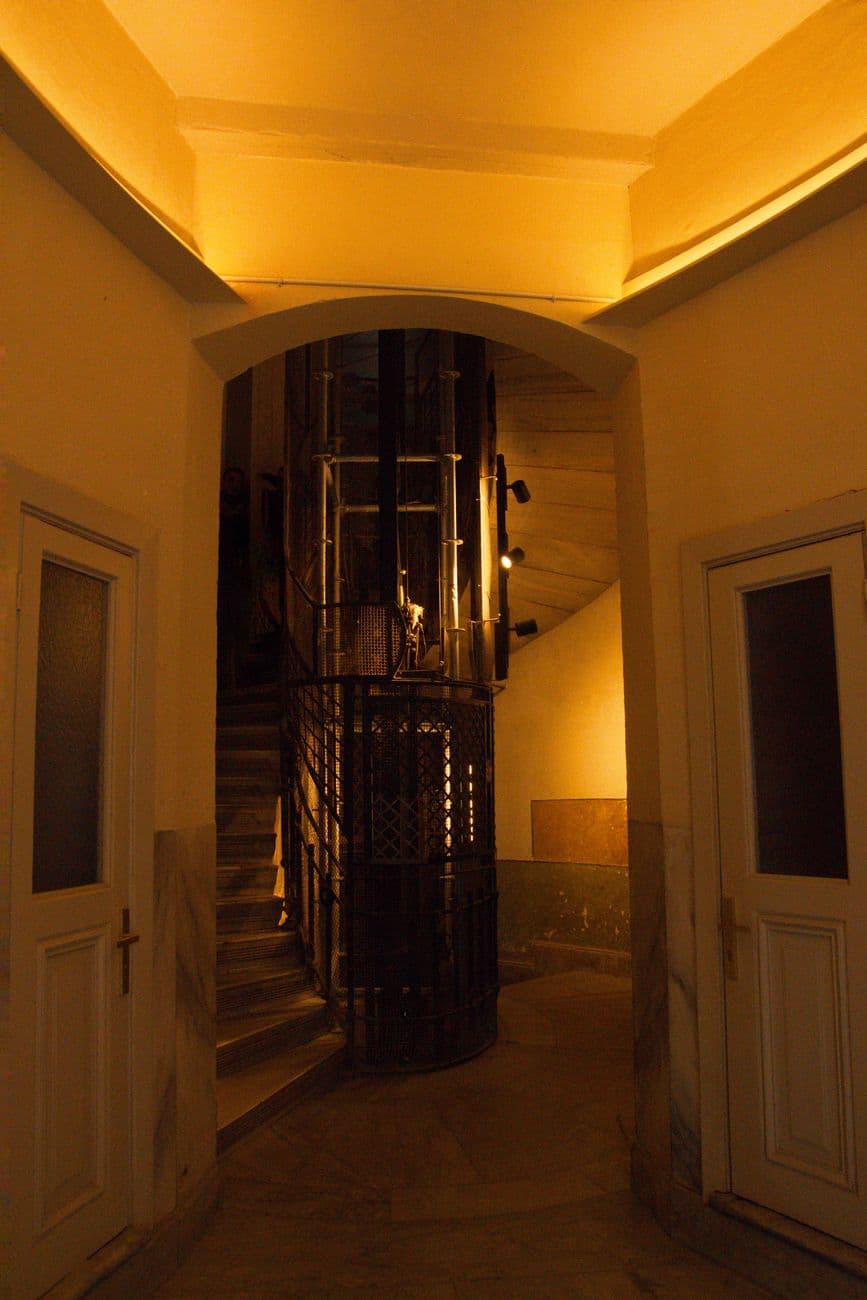 Dimly lit vintage staircase with metal lift, encased by rounded walls and doors.