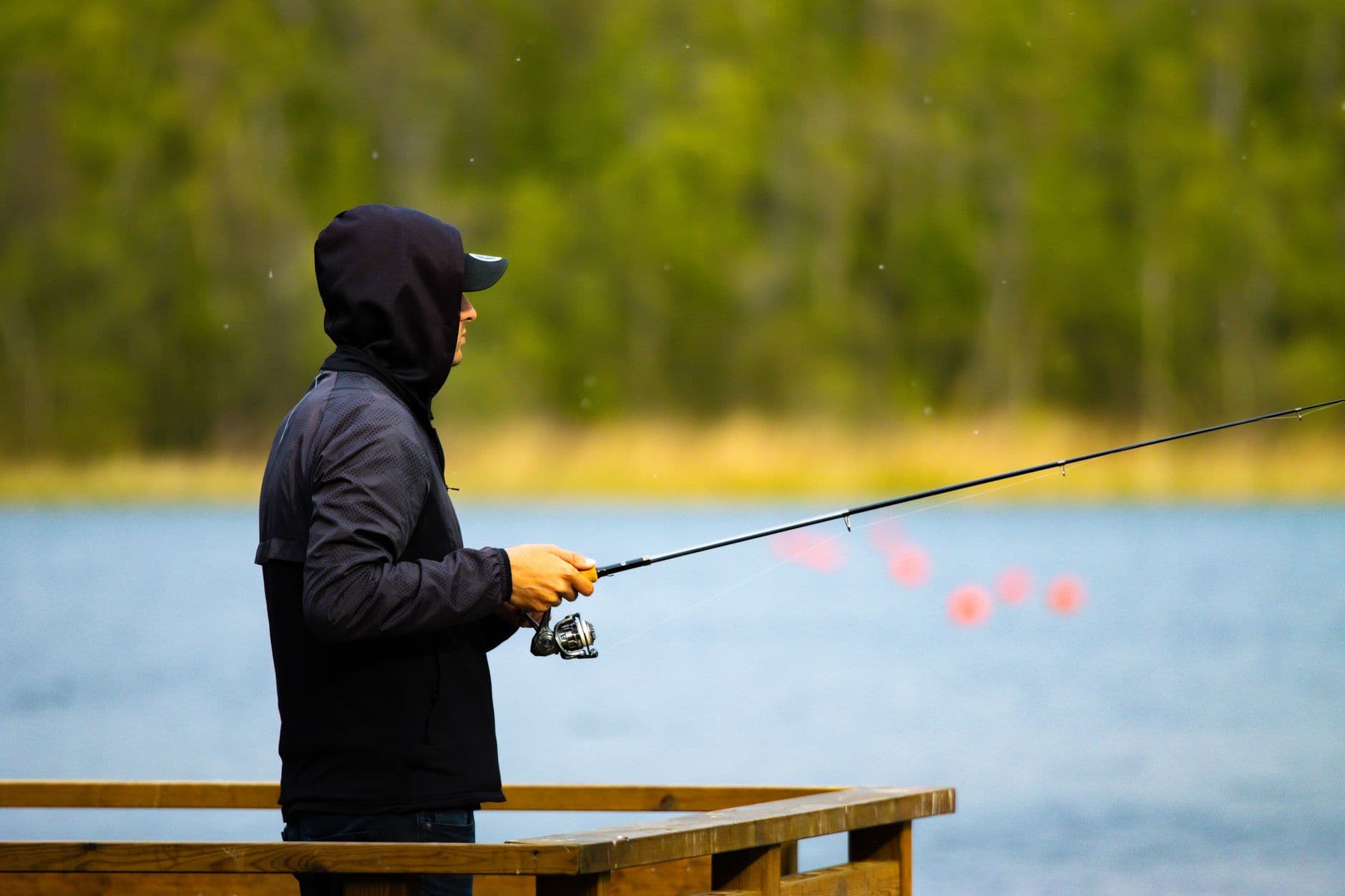 Man in a hoodie fishing on a wooden dock by a lake in Jönköping, Sweden.