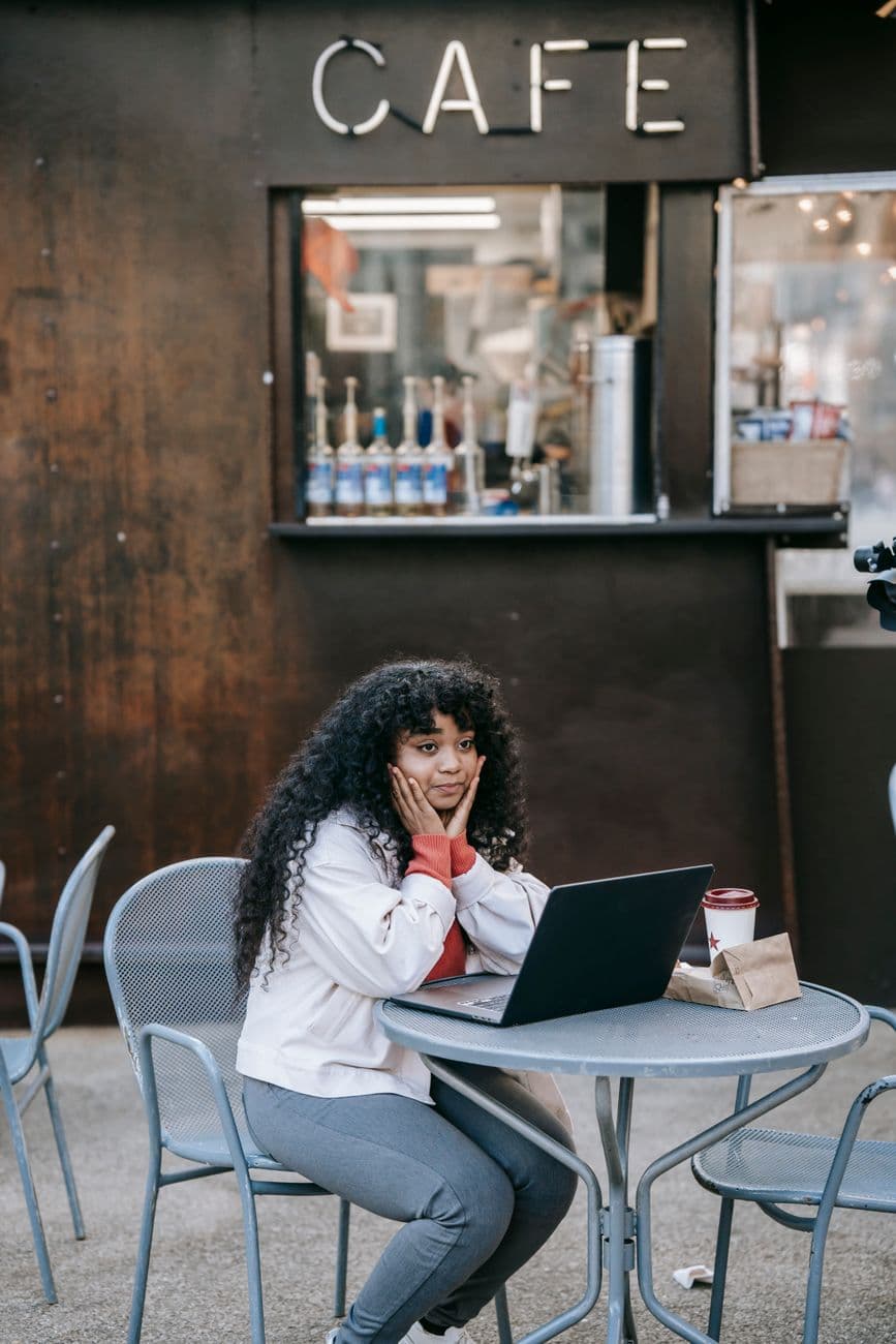 Positive African American female wearing casual clothes sitting at outdoor cafe table with netbook and takeaway coffee while touching face and looking away in pleasant thoughts
