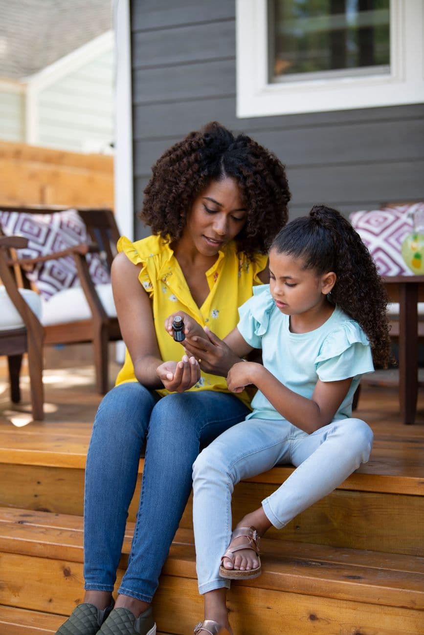 A mother and daughter sitting on wooden steps applying essential oil, enjoying a sunny day.