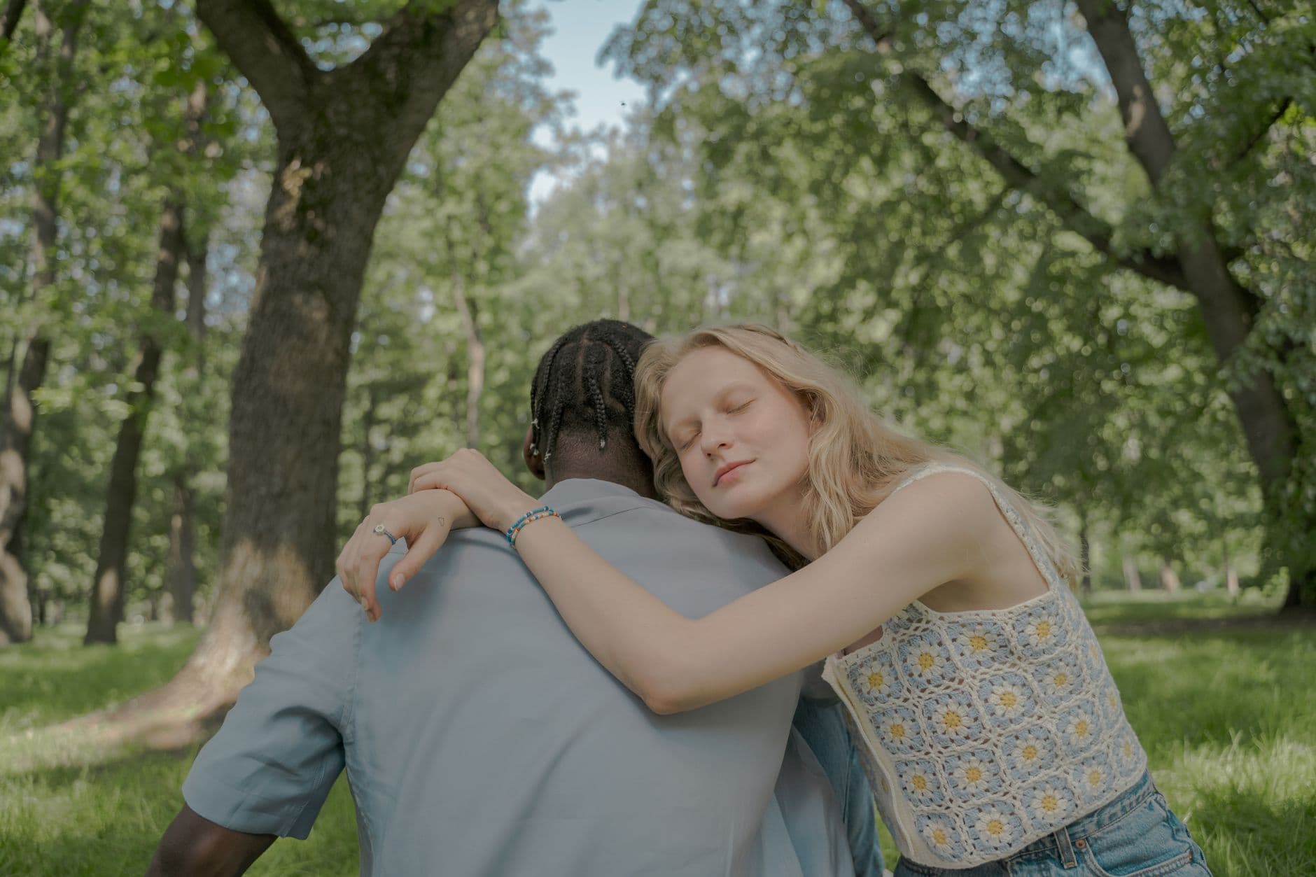 A young couple enjoying a warm hug while sitting outdoors in a lush green park during a sunny summer day.