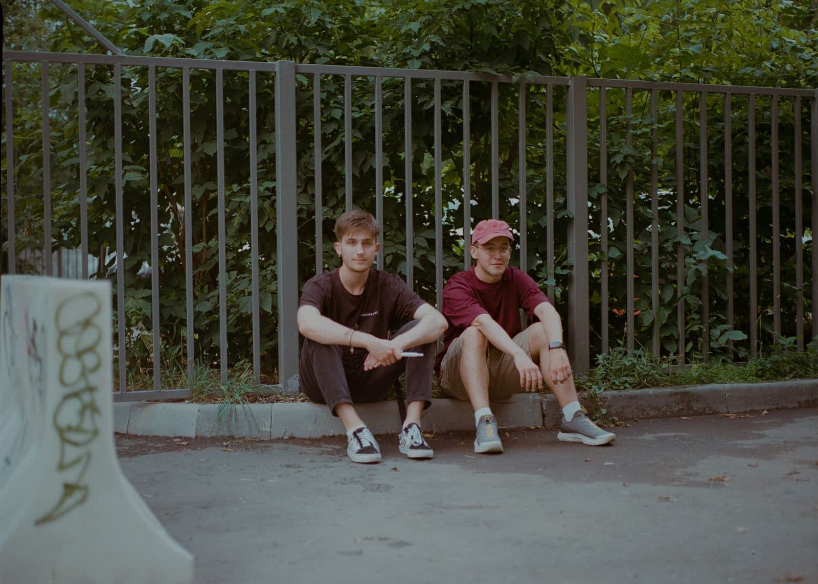 Two young men sitting by a fence in a Moscow park, enjoying a relaxed moment together.