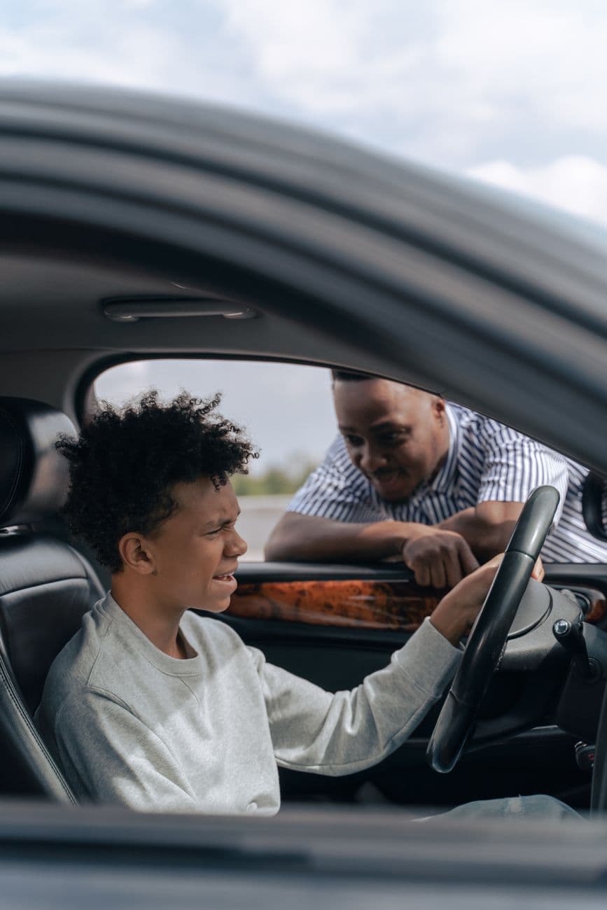 Father instructs son in car driving lesson inside a vehicle on a sunny day.