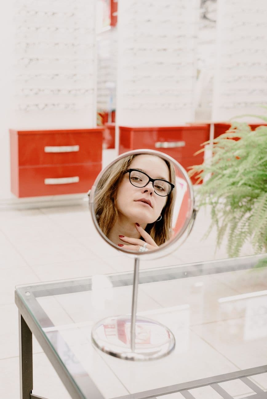 Crop female client reflecting in mirror while choosing glasses for correcting eyesight in eyewear store
