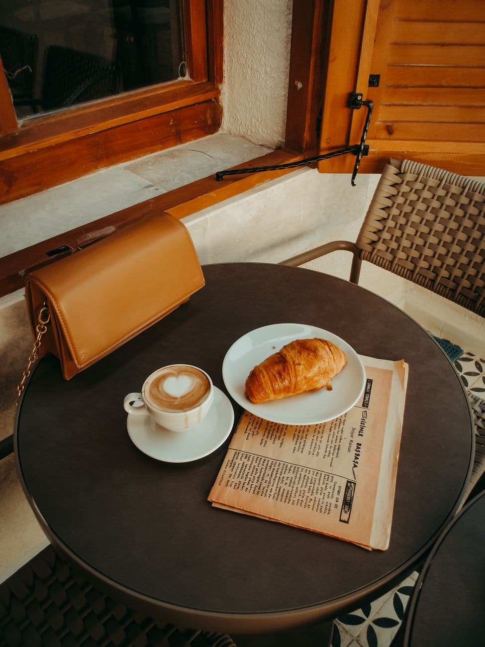 A chic cafe scene with a cappuccino, croissant, and handbag on a round table.