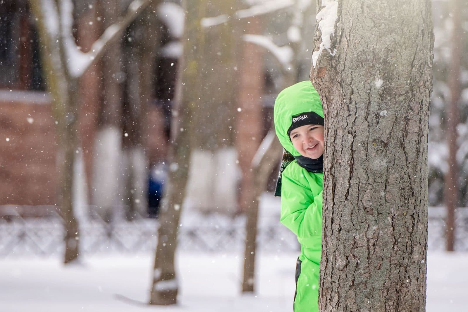 Smiling child in green winter outfit hiding behind a tree during snowfall.