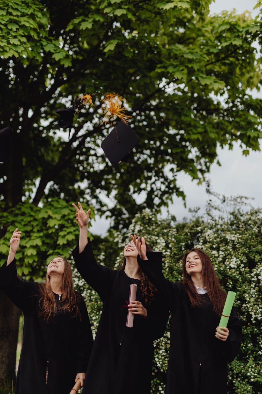 Three graduates in gowns throwing caps high, celebrating success outdoors.