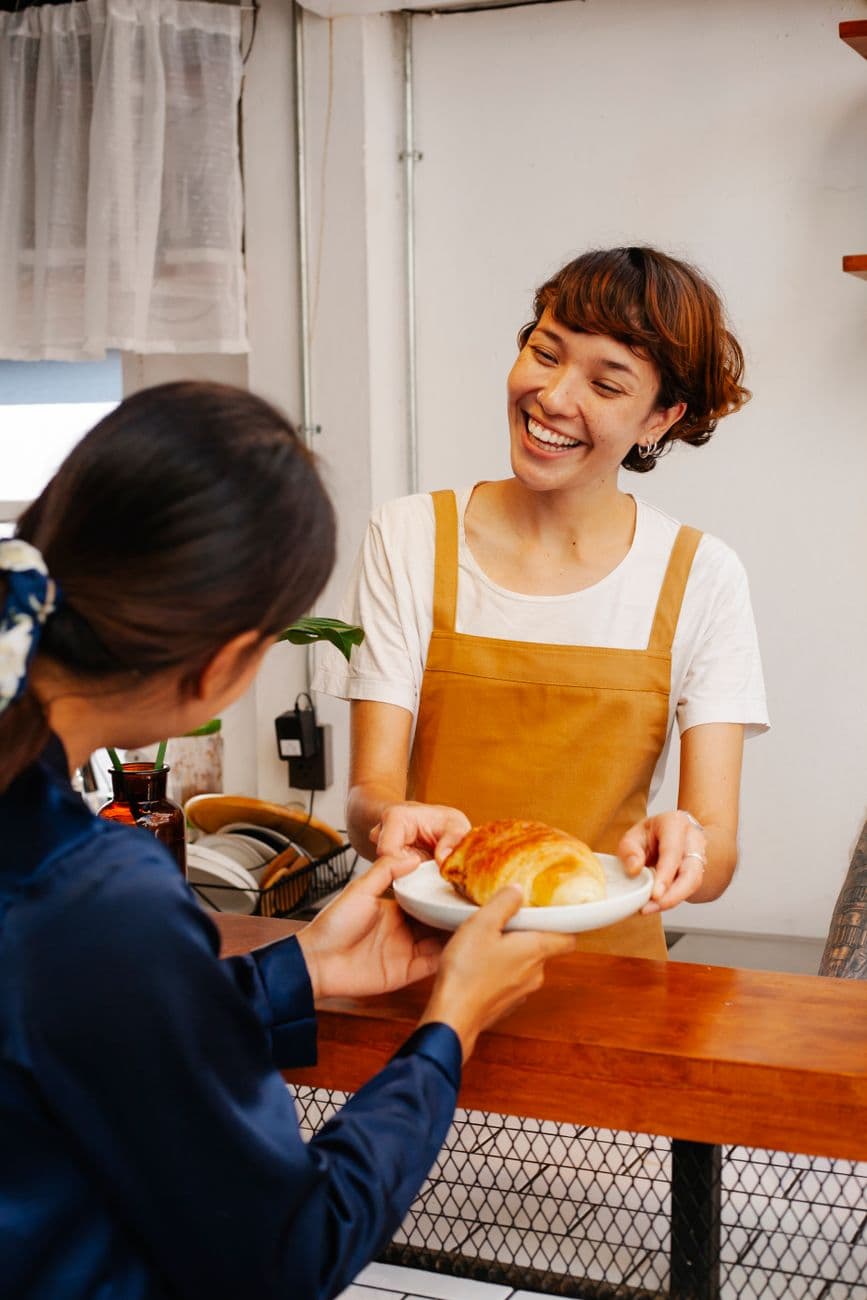 Happy female worker in apron passing appetizing puff to anonymous colleague over counter in cafe