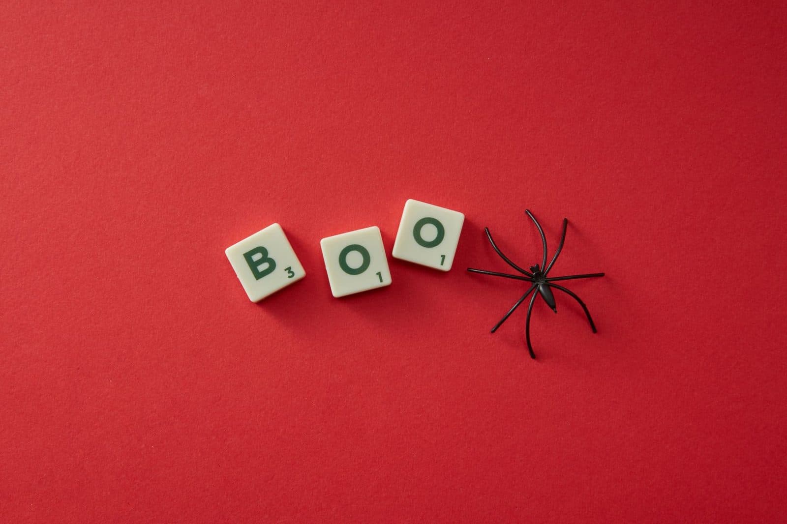 Scrabble tiles spelling 'BOO' with a black plastic spider on a red background, perfect for Halloween themes.