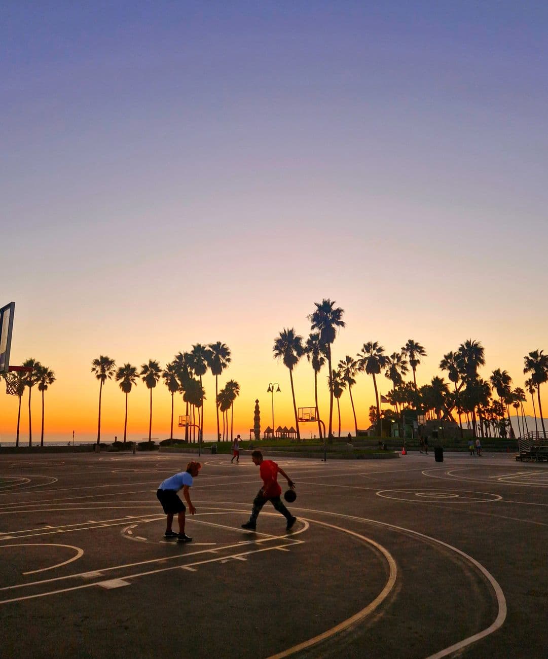 Two people playing basketball at sunset on Venice Beach, Los Angeles, surrounded by palm trees.