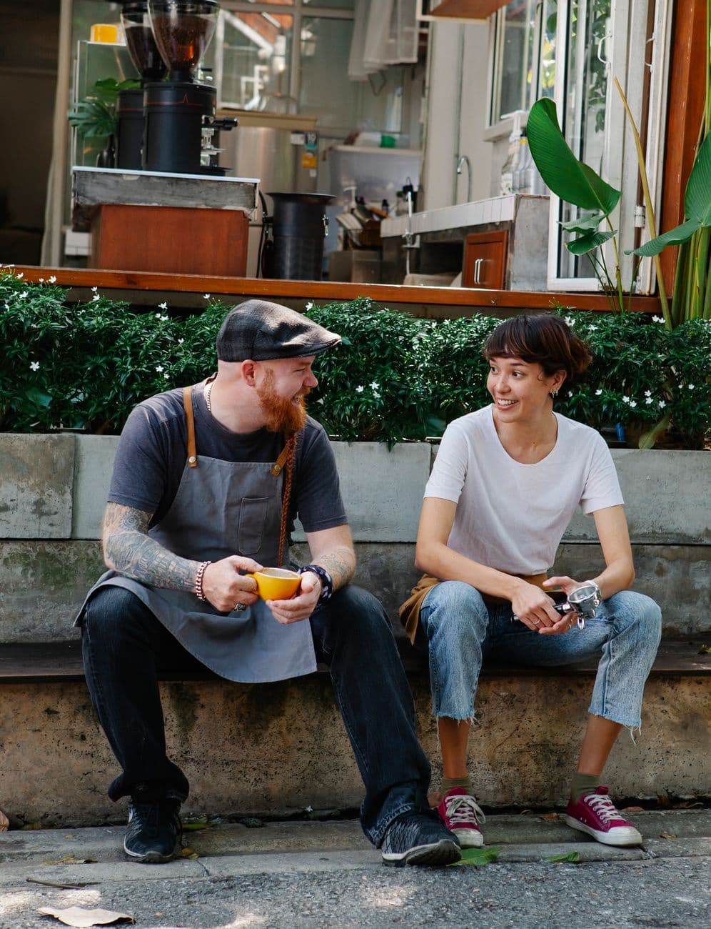 Full length of smiling couple of barista coworkers in aprons and casual outfit sitting in street near cafeteria and plants while talking with cup of drink and portafilter in daytime