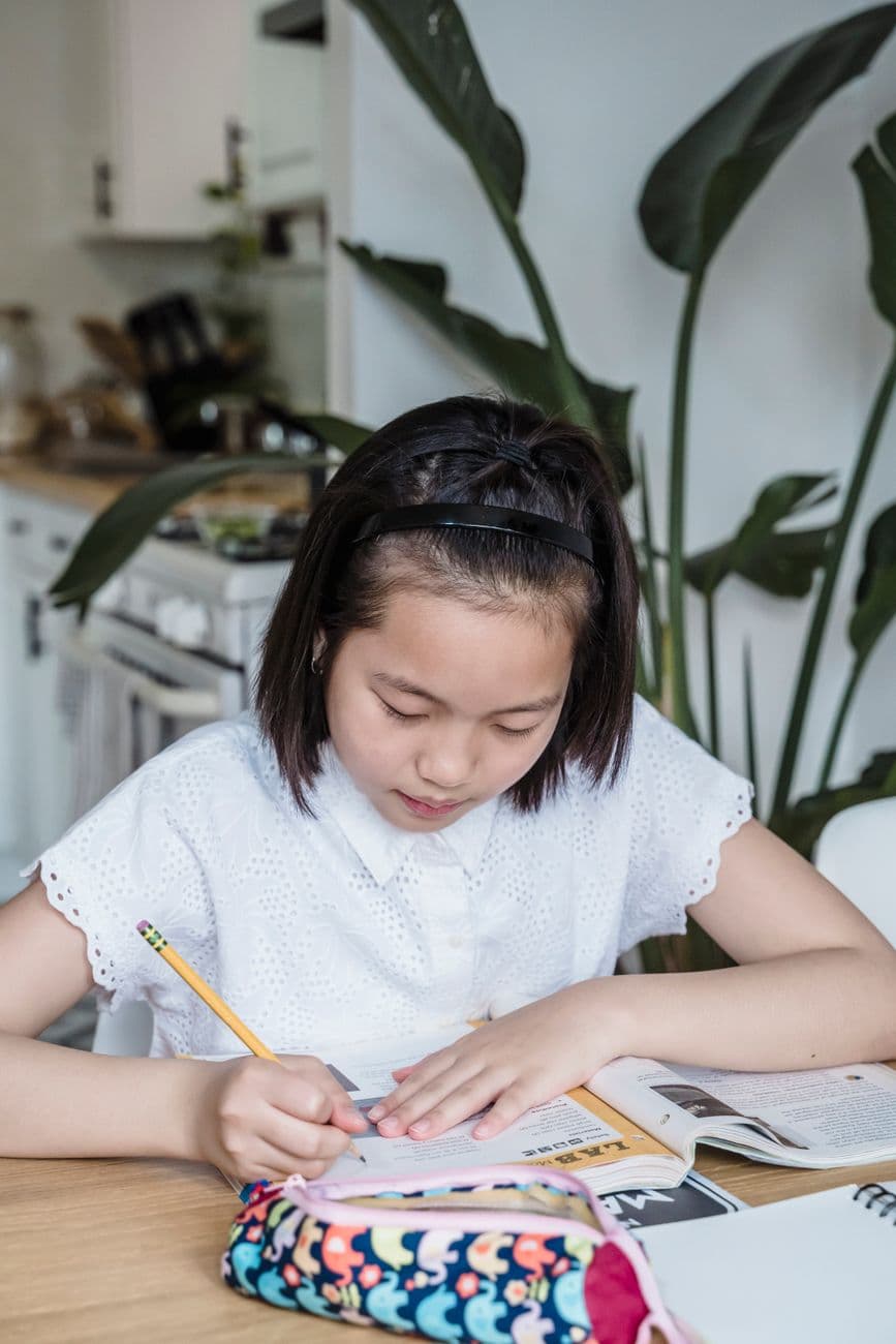 A young girl writing on paper while studying at a wooden table in a modern kitchen setting.