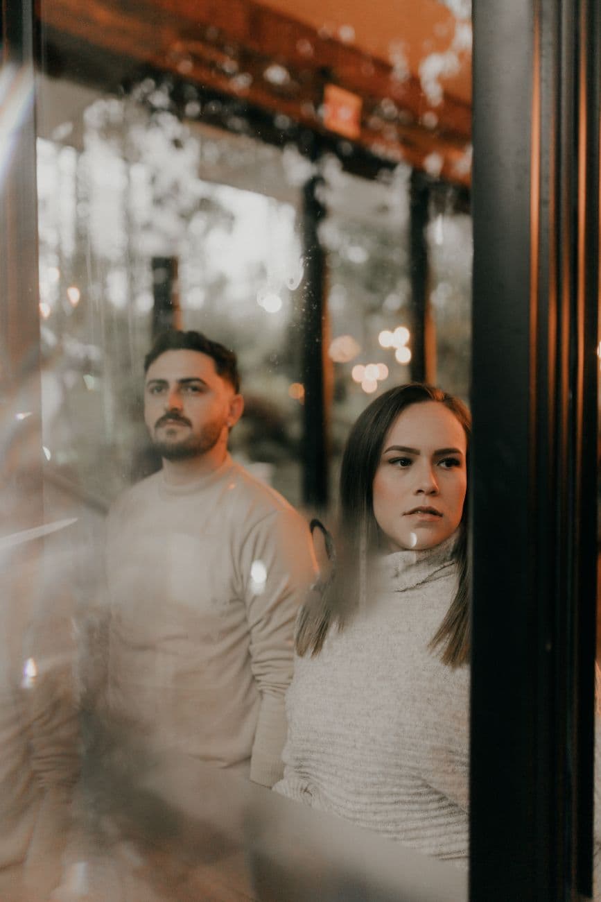 A thoughtful couple stands by a window with reflections, capturing a serene indoor moment.