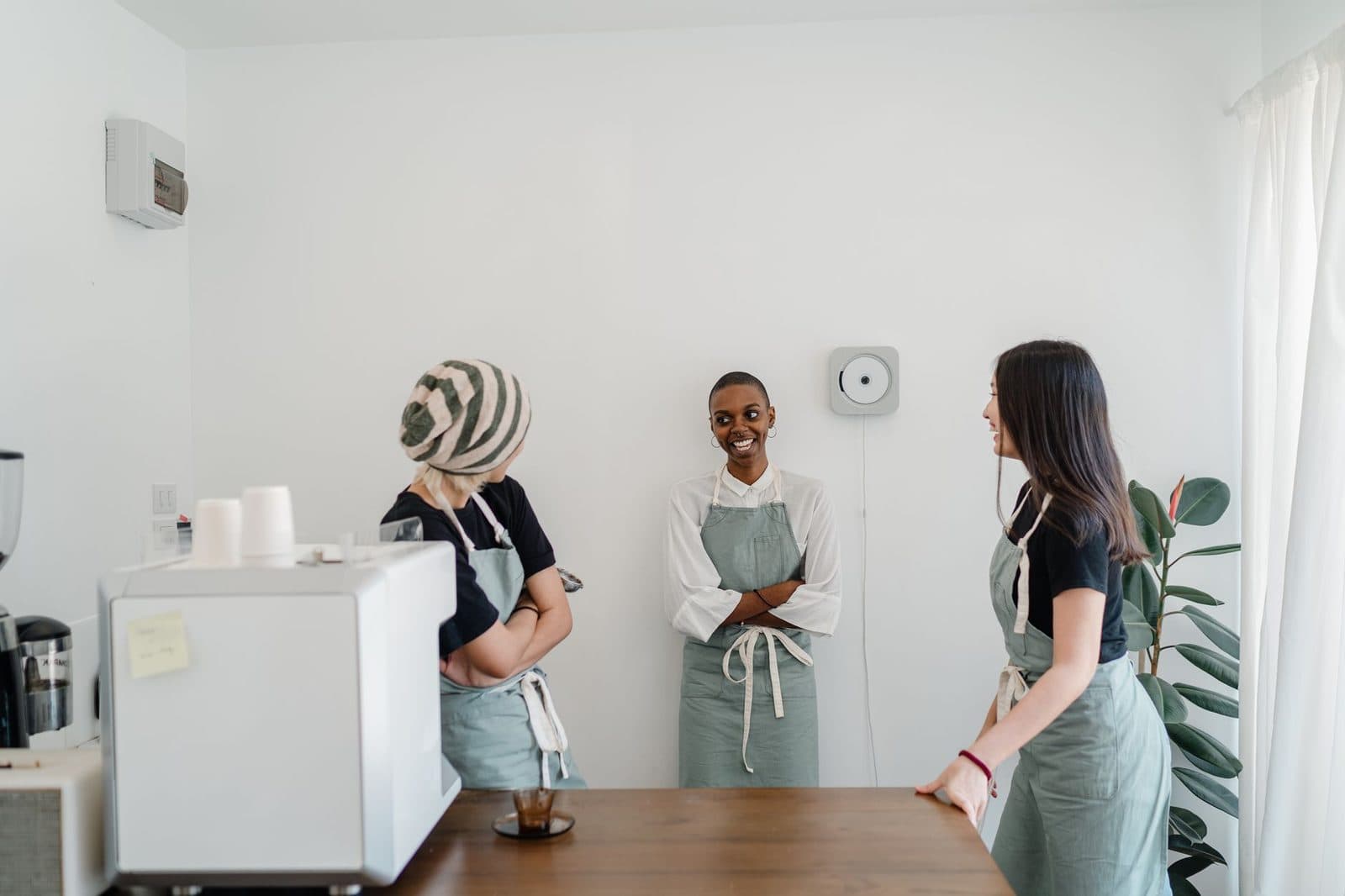 Three female baristas chatting and smiling in a modern, bright cafe setting.