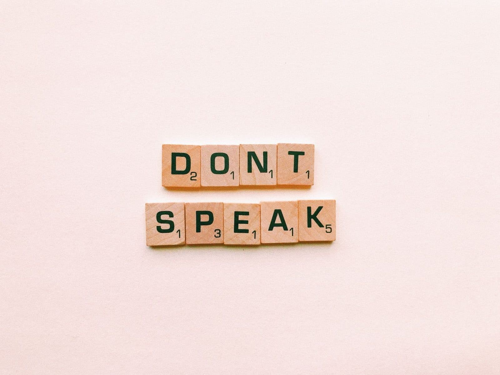 Wooden Scrabble tiles spelling 'Don't Speak' on a pink background, emphasizing silence.