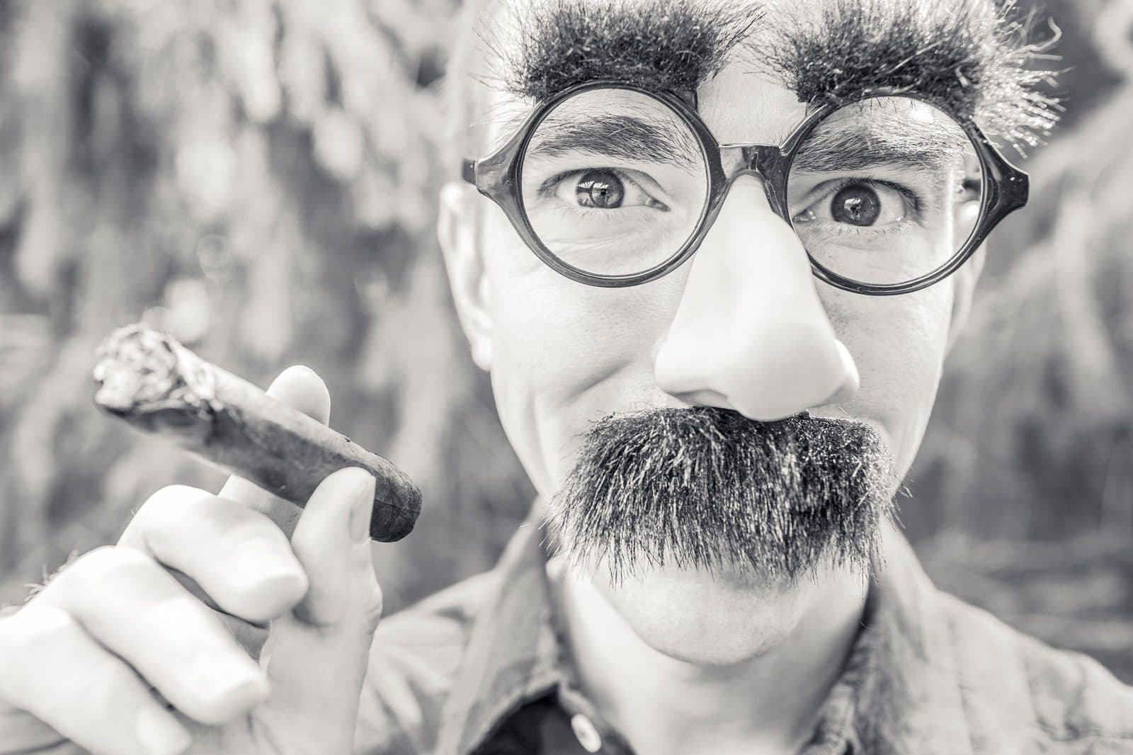 Silly portrait of a man wearing costume glasses and mustache, holding a cigar.