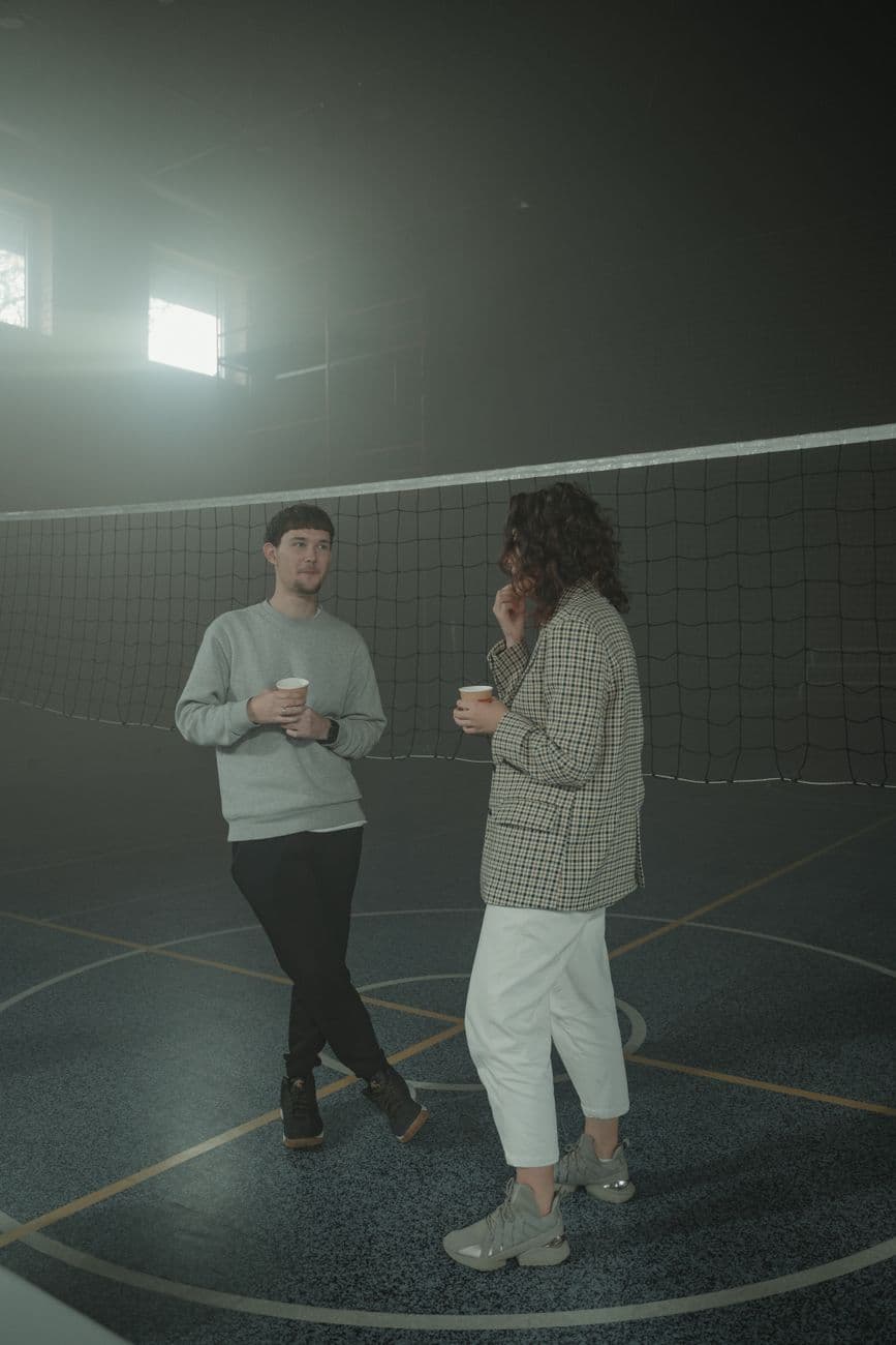 A man and woman chatting casually over coffee in an indoor sports hall with a volleyball net.