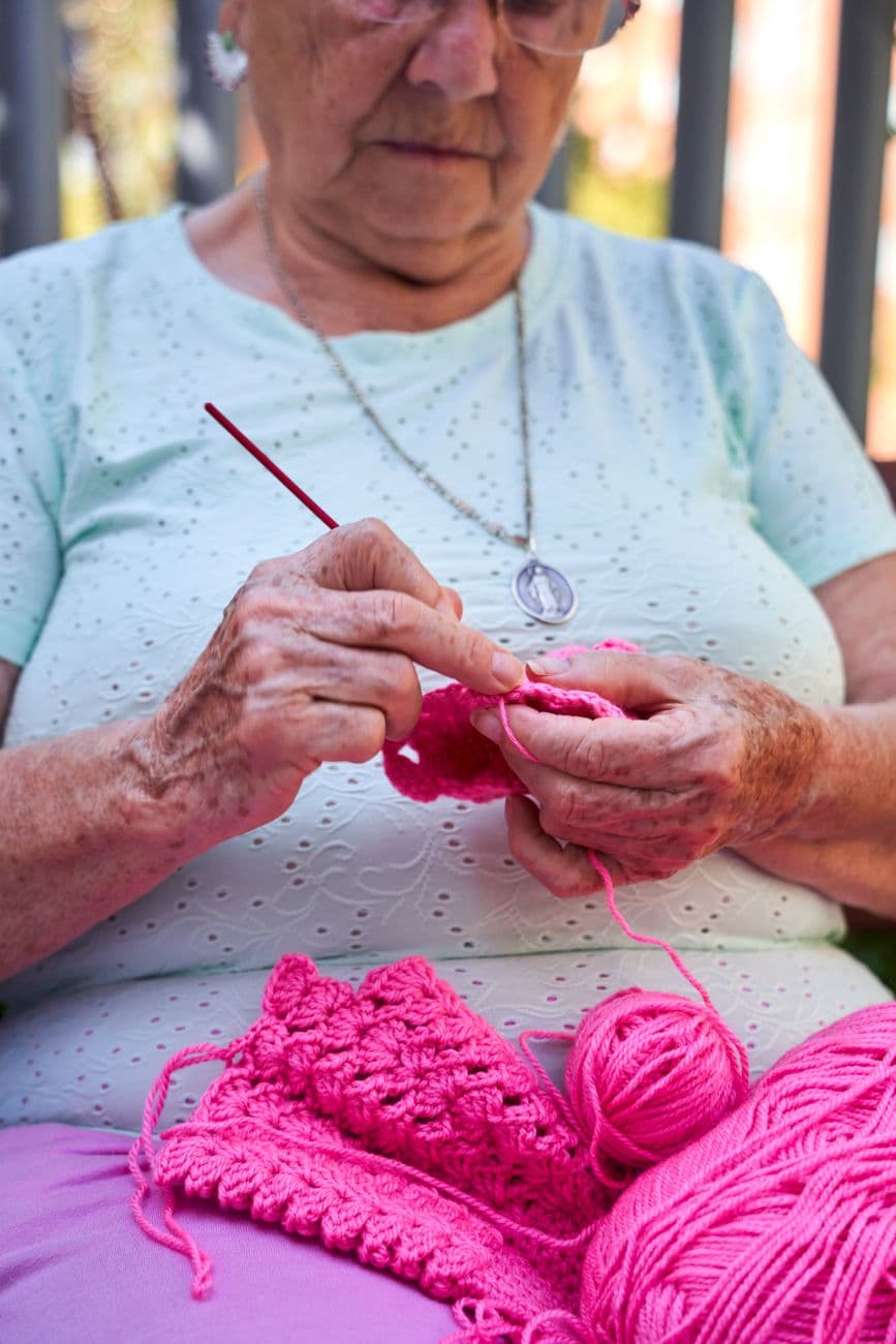 Close-up of a senior woman crocheting pink yarn with focus on hands and needles.