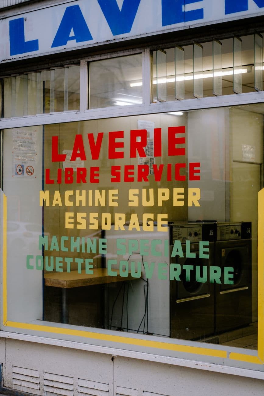 Front view of a self-service laundromat window with colorful text and washing machines inside.