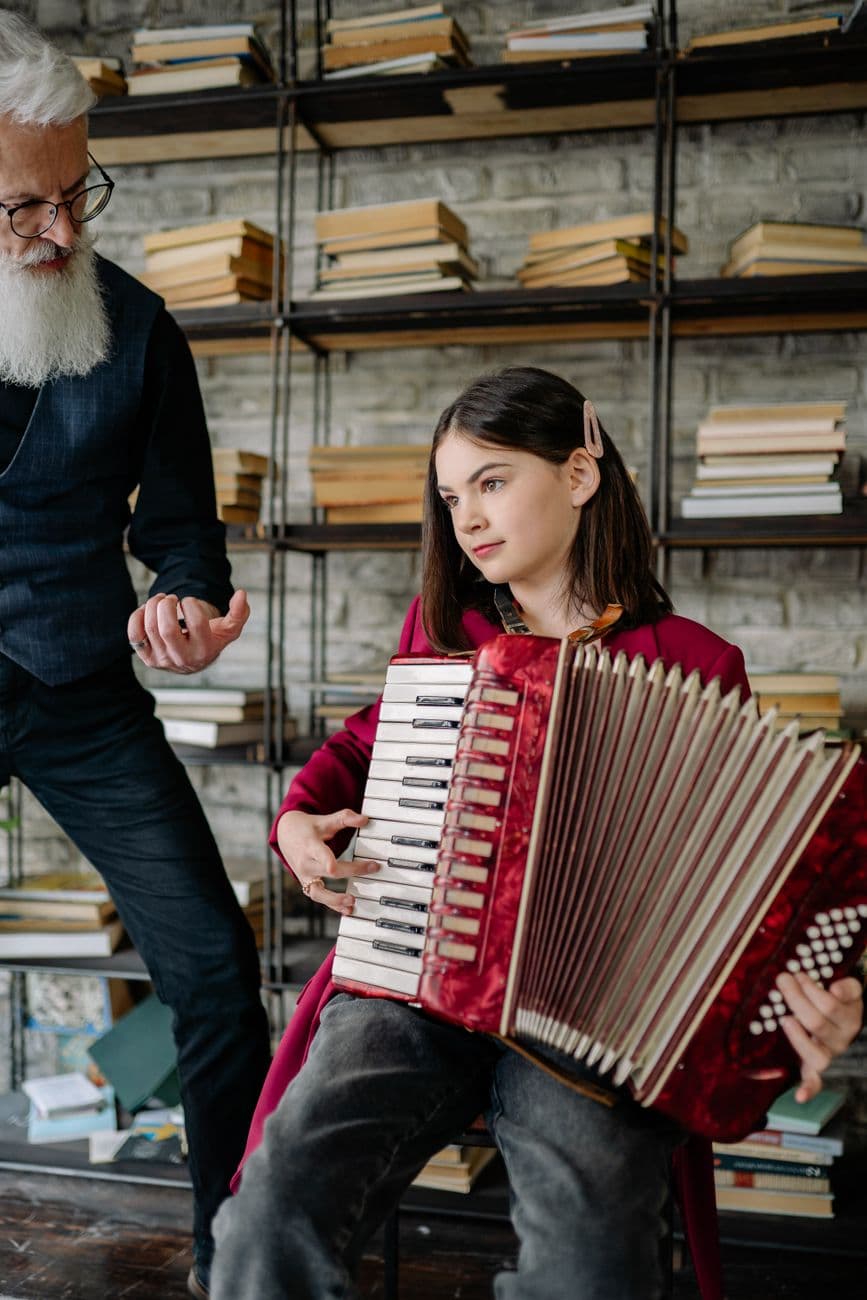 A young girl learning to play accordion under a teacher's guidance in a cozy library setting.