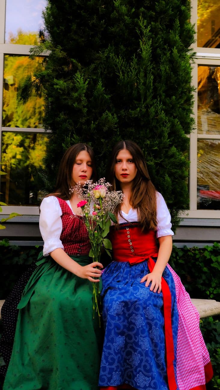 Two Austrian women in vibrant traditional dirndls holding flowers, seated outdoors in Graz, Austria.