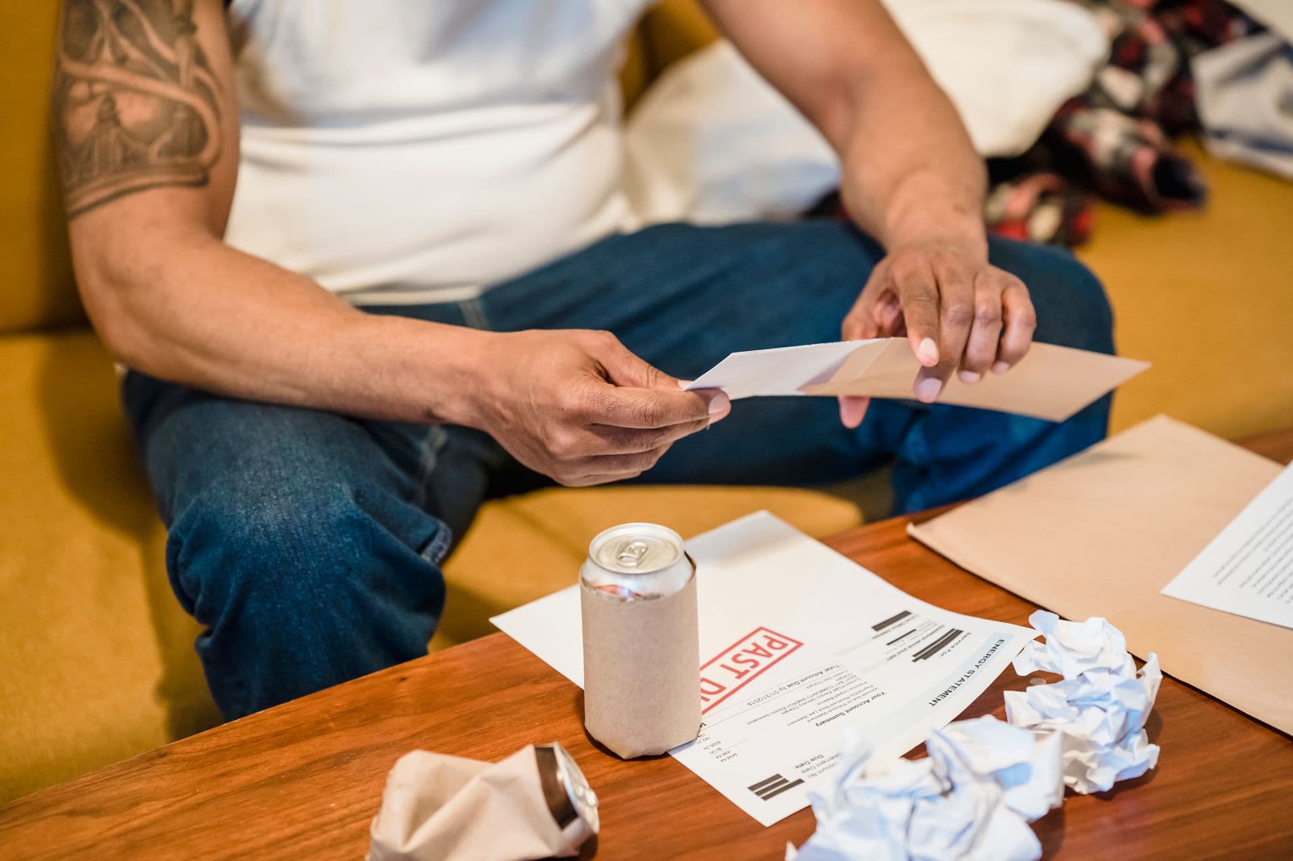 A man inspects his mail while seated on a couch, envelopes and papers scattered.